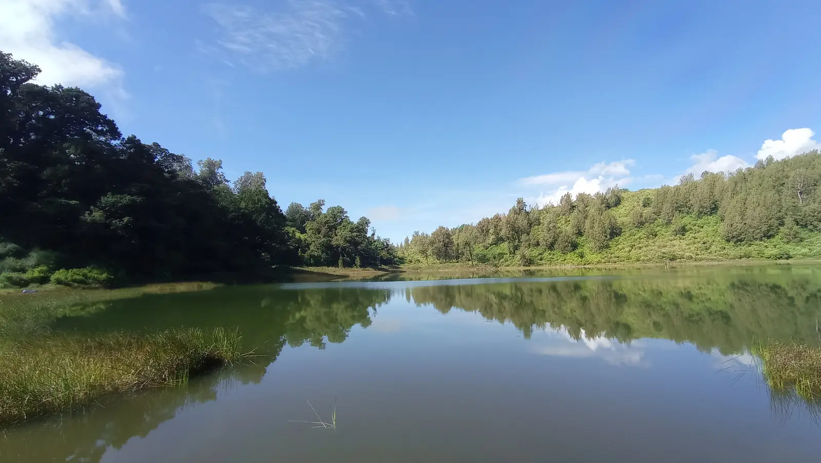 Danau Ranu Regulo yang tenang memantulkan bukit hijau dan langit di kaki Gunung Semeru